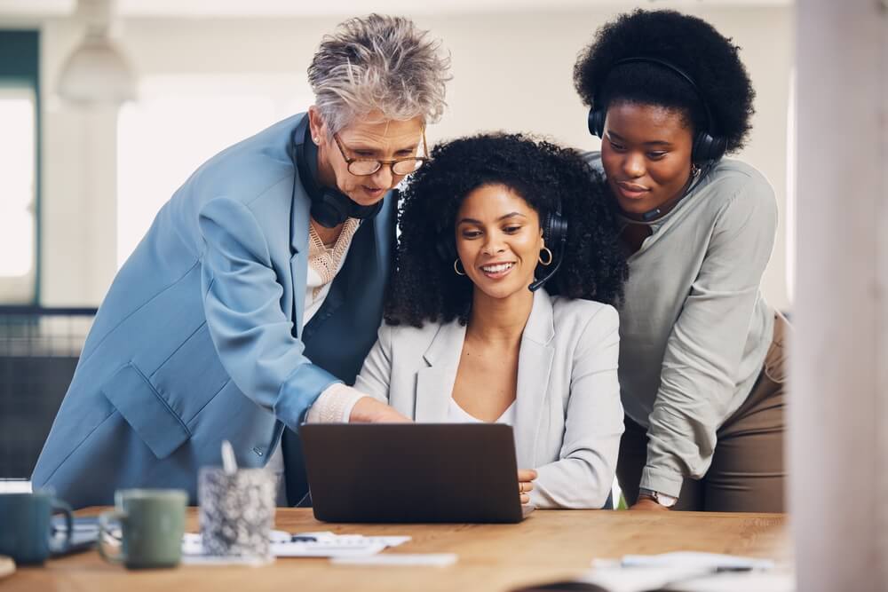 A imagem mostra três mulheres. A da esquerda é branca, enquanto as outras duas são negras. A do meio está mexendo em um notebook, enquanto as outras duas mulheres observam, como se explicassem algo.