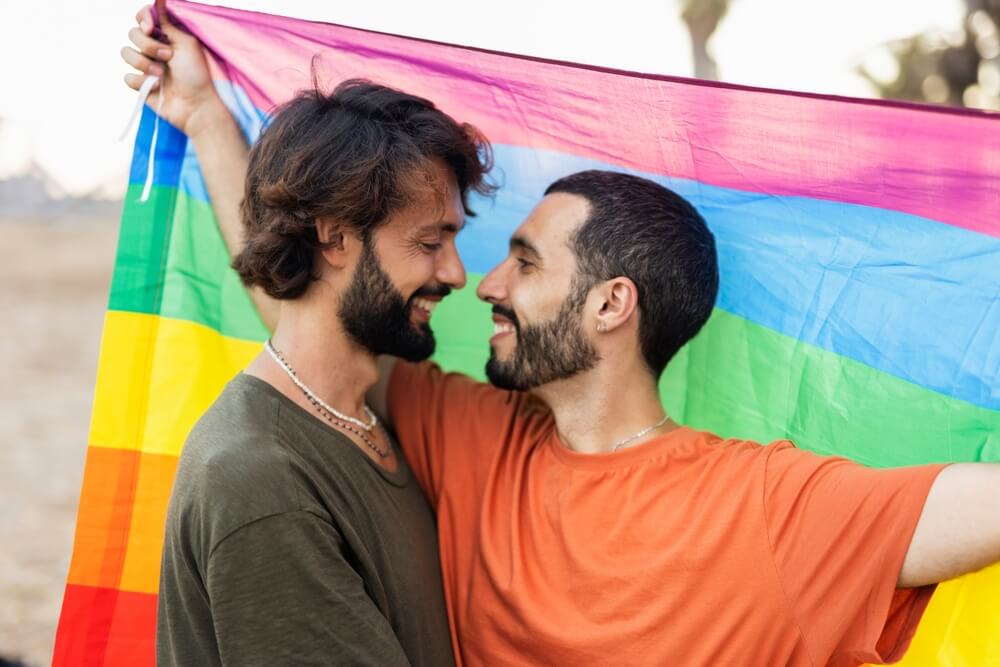 Imagem de dois homens. Eles são namorados e estão segurando, ao fundo, a bandeira LGBTQIA+.