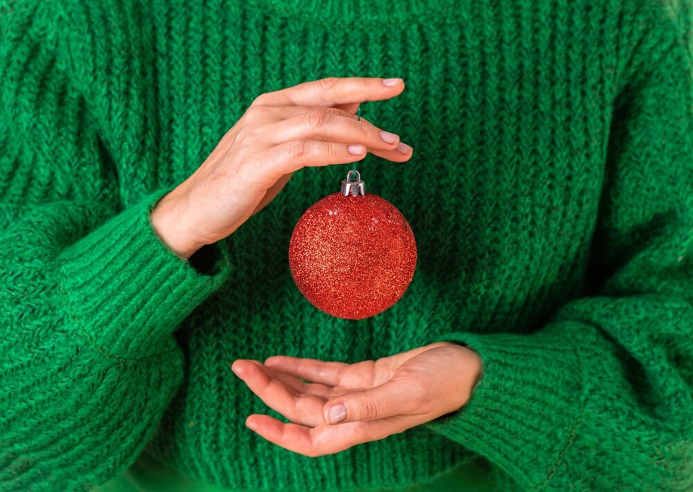 Imagem de uma mulher. O foco está nas mãos dela. Ela está segurando uma bola vermelha de Natal, e utiliza uma blusa de frio verde.