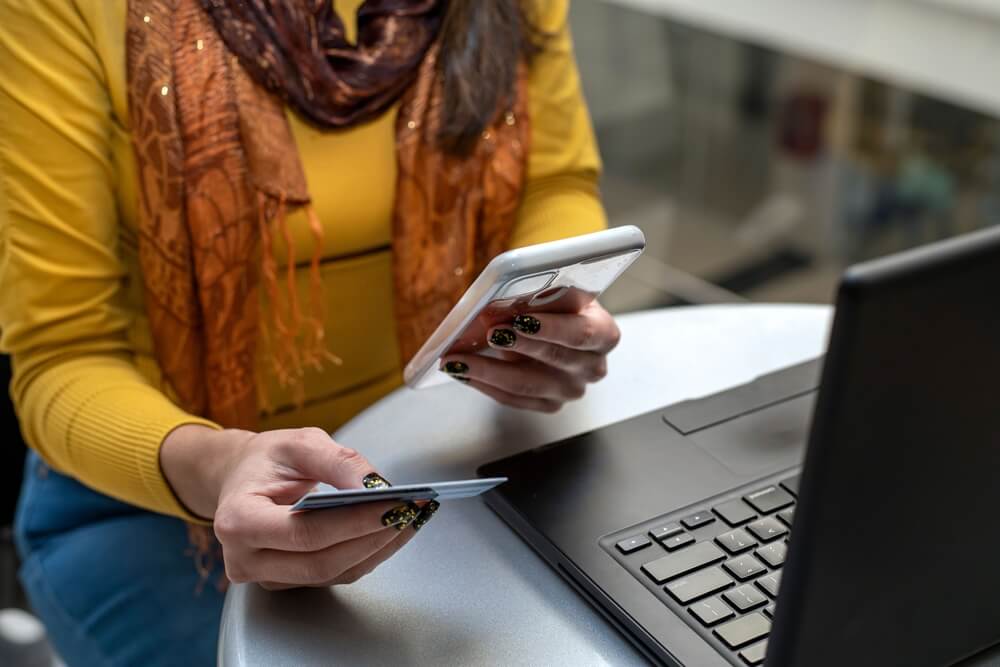 Imagem de uma mulher sentada em frente ao computador. Ela está com o cartão de crédito em uma mão e o celular em outra.