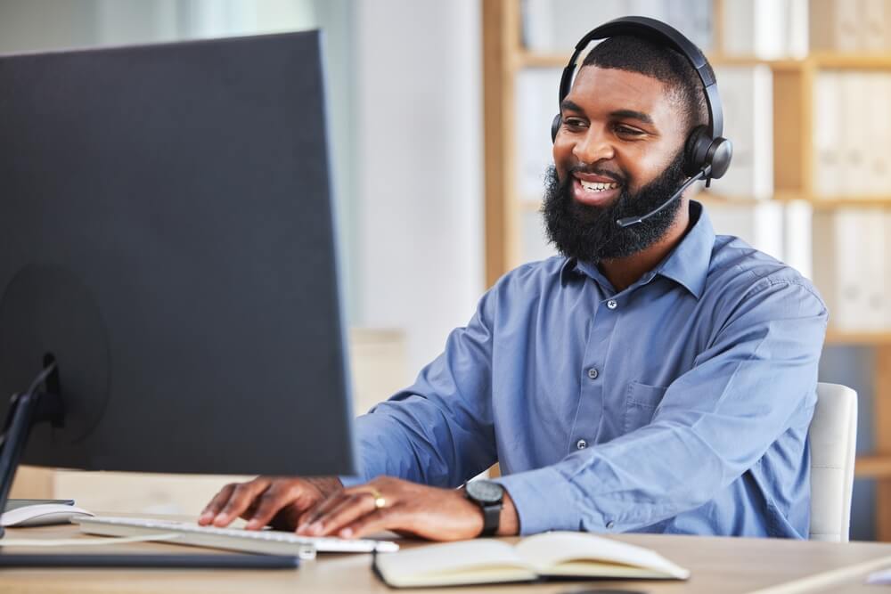 Imagem de um homem negro. Ele está mexendo em um computador e com um fone de ouvindo, prestando serviços de atendimento ao cliente.
