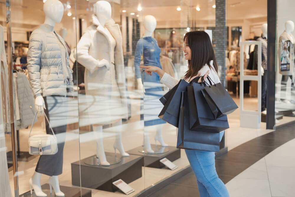 Uma mulher branca, de cabelos escuros, olhando a vitrine de uma loja. Ela carrega diversas sacolas, pois fez compras durante a Black Friday.
