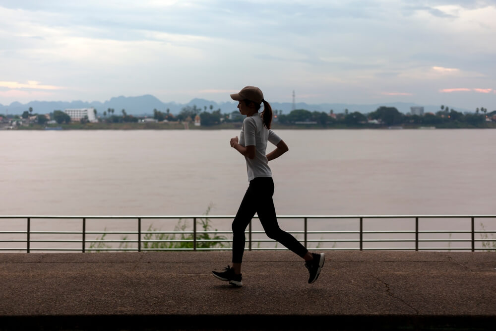 Imagem de uma mulher praticando exercício físico na frente de um rio. Ela está correndo.