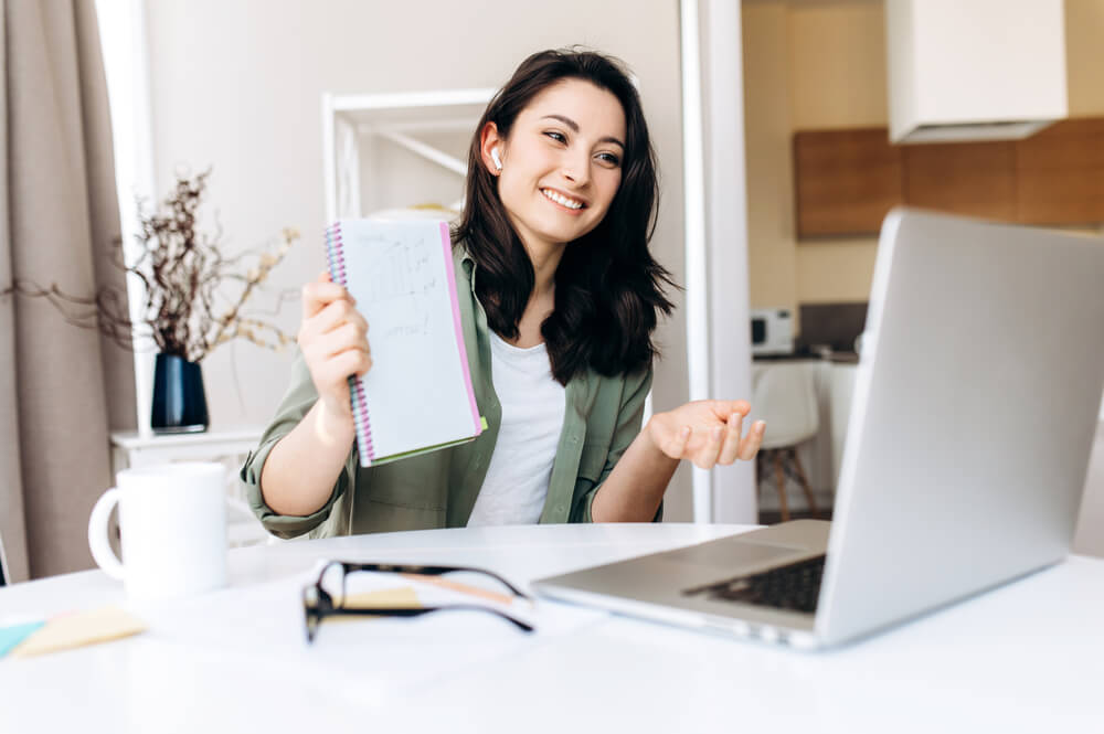 Uma mulher de cabelos pretos em frente a um computador. Ela segura um caderno, como se estivesse ensinando algo a alguém por meio da internet.