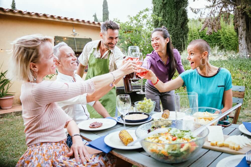 Várias pessoas ao redor de uma mesa, brindando com vinho. Elas parecem estar contentes e estão na área de uma casa, em meio a um jardim.