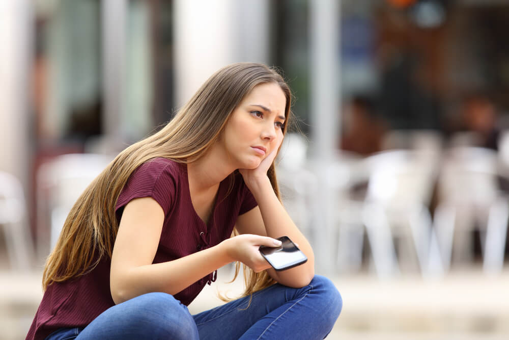 Imagem de uma mulher sentada em uma calçada. Ela veste um camiseta roxa, tem os cabelos loiros e segura um celular. Parece estar descontente com algo.