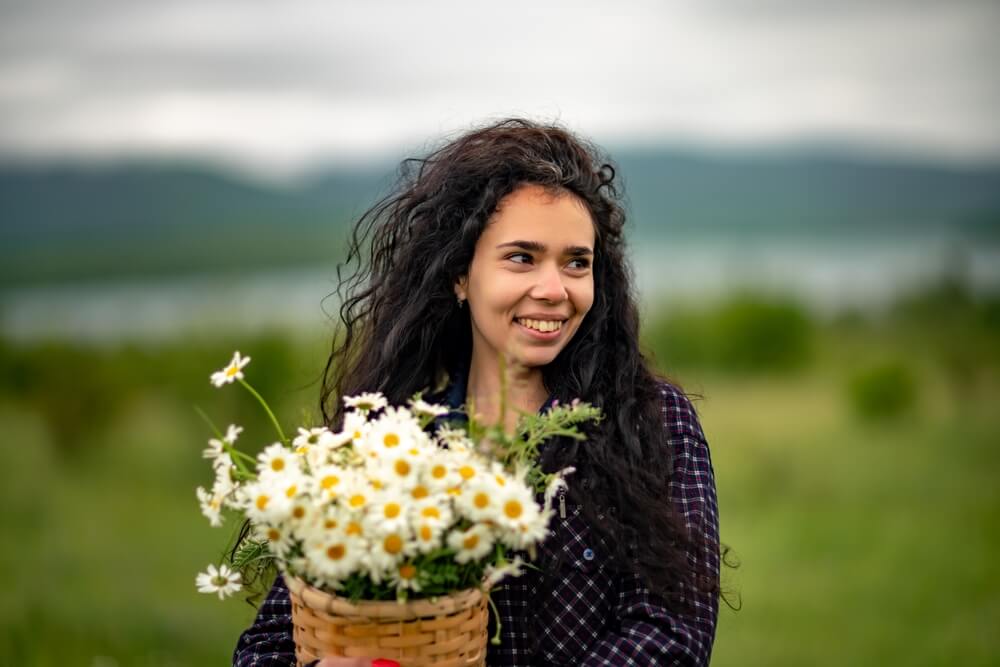Mulher de cabelos pretos. Ela segura uma cesta cheia de flores brancas. Parecem margaridas. Ela está sorrindo, com uma aparência de contente.