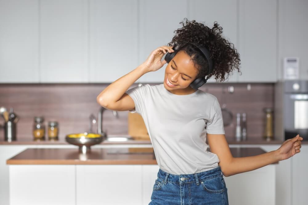 Imagem de uma mulher negra dançando na cozinha. Ela tem cabelos encaracolados e utiliza fones de ouvido.