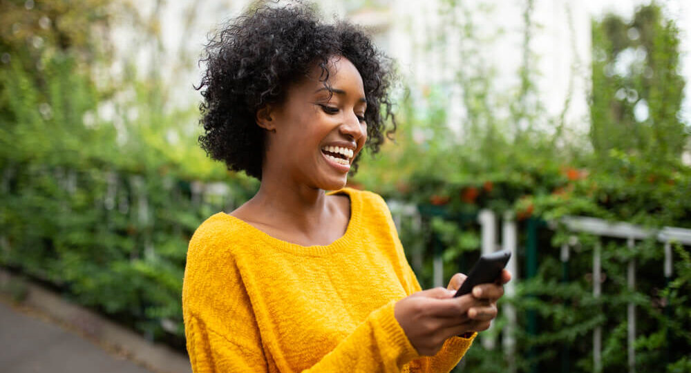 Imagem de uma mulher negra, usando uma blusa de manga comprida amarela, enquanto mexe no celular. Ela está sorrindo.