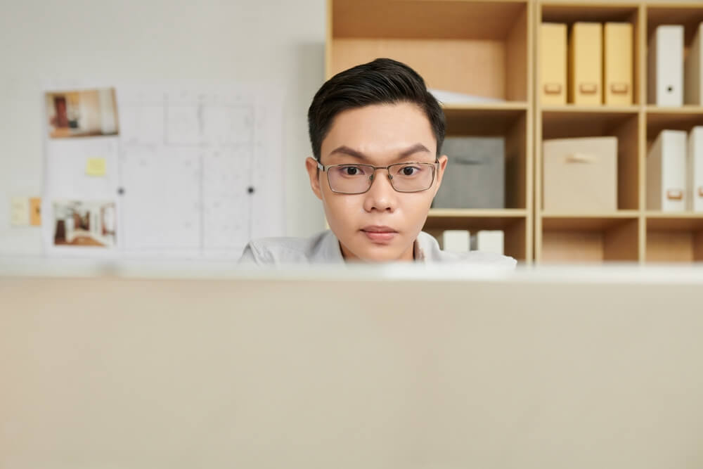 Imagem de um menino branco sentado em frente a um computador. Ele tem traços orientais e usa óculos.