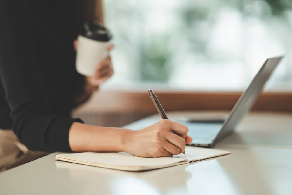 Imagem de uma mulher escrevendo em um caderno. Ao lado, há um notebook. Ela está segurando um copo térmico de café.