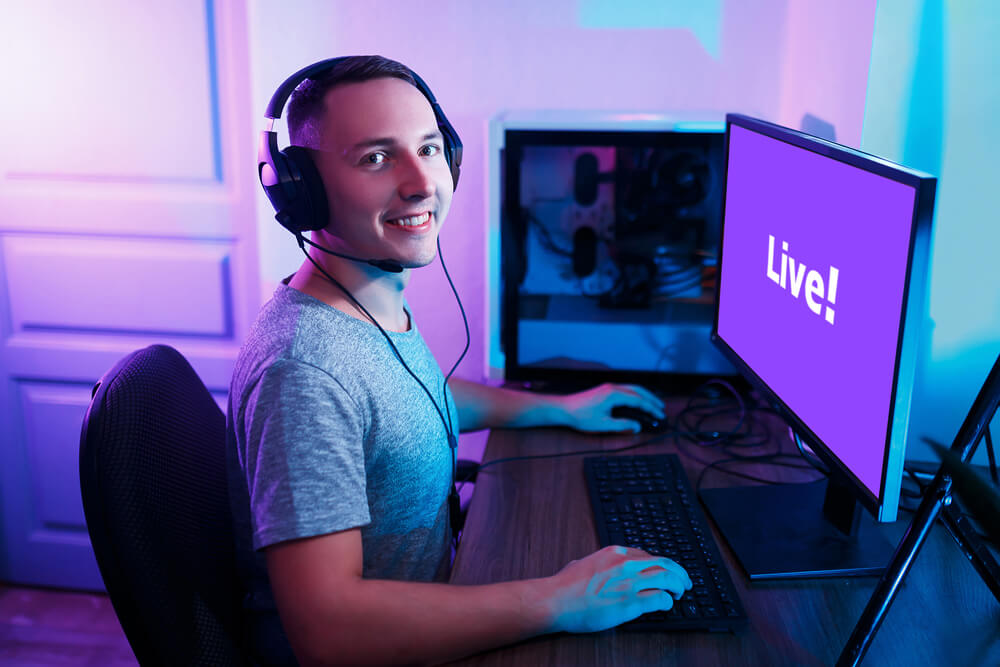 Imagem de um homem branco com um fone de ouvido em frente a um computador. Ele está fazendo uma live na Twitch, uma plataforma de streaming roxa que também pode ser considerada uma das mídias alternativas.