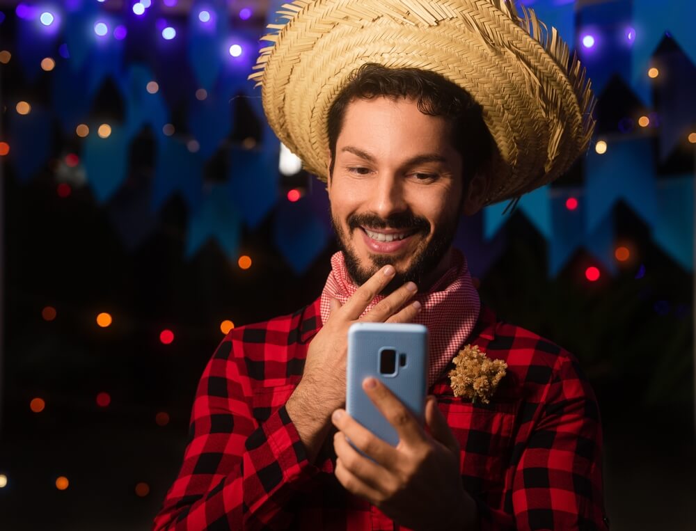 Imagem de um homem branco e barbudo segurando um celular. Ele veste uma camisa xadrez vermelha e está com um chapéu de palha na cabeça.